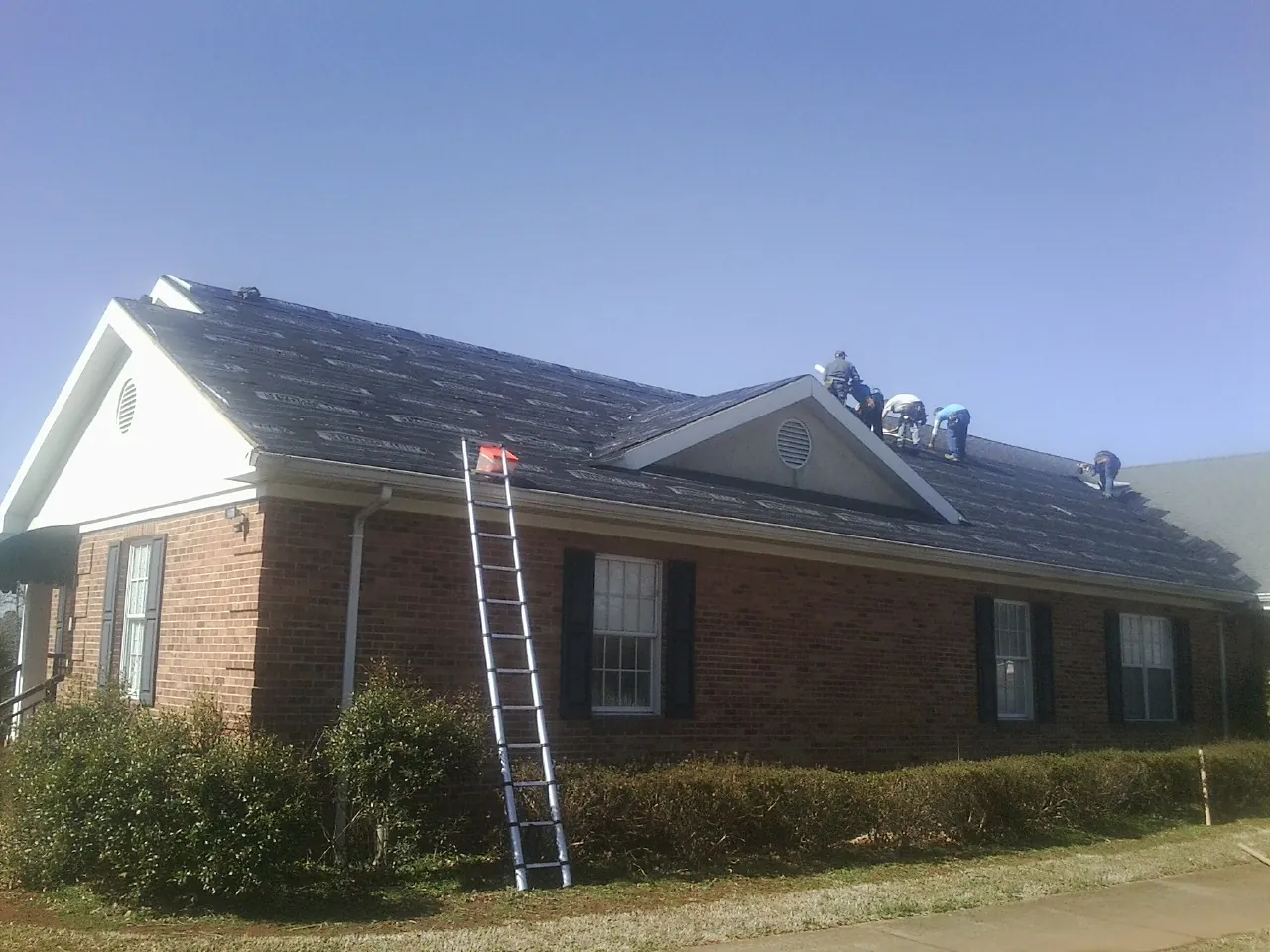 Crew tearing shingles off a brick ranch roof with a ladder set — Metal Roof Repair work by our crew in Collins